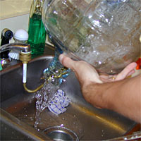 Person pouring water from a large bottle into a kitchen sink surrounded by various items.